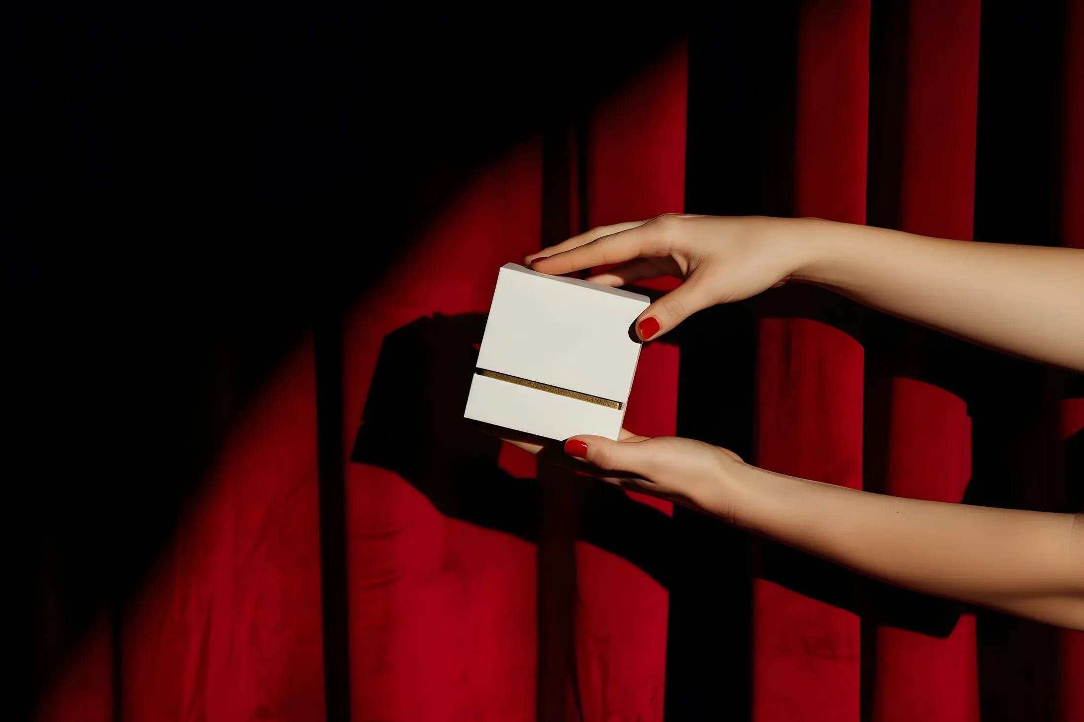 Two hands holding a white gift box against a red velvet curtain